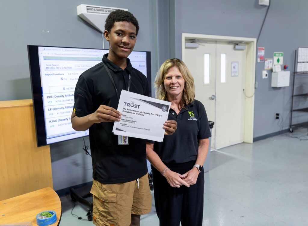 A young man holds a certificate while standing next to a smiling woman in a classroom. A screen with text is behind them, and both are dressed in black shirts.