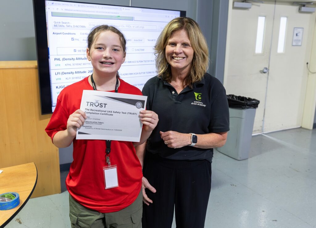 A young person in a red shirt holds a certificate and stands beside an adult woman in a black shirt. They are smiling indoors, with a large digital screen in the background displaying text.
