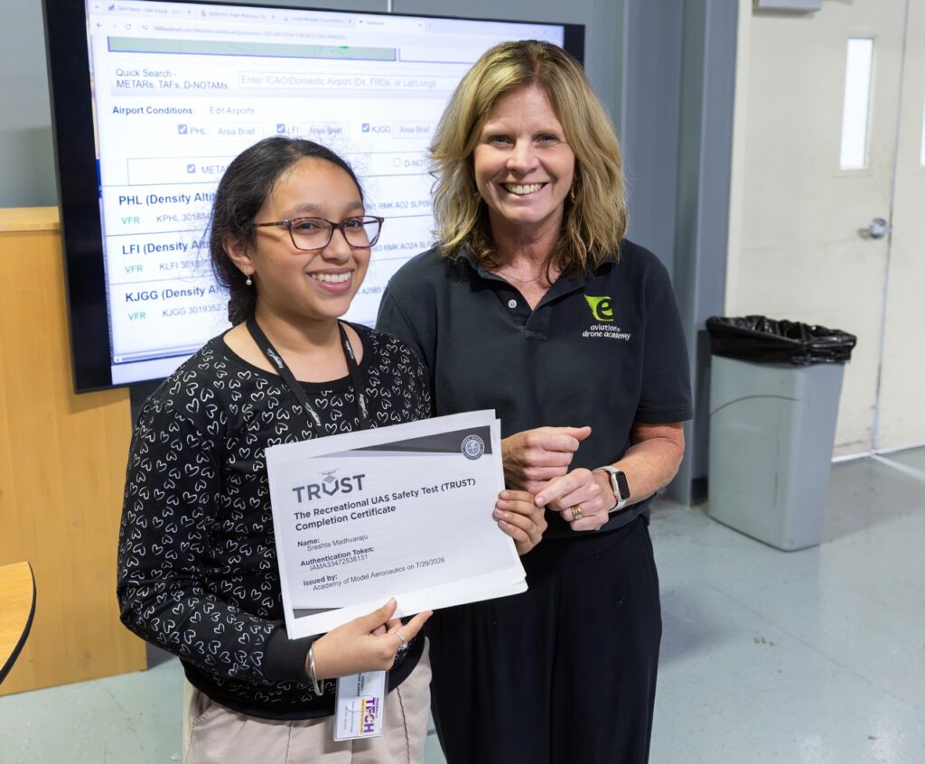 Two women smiling indoors; one holds a printed TRUST certification. A trash can, door, and computer monitor displaying flight information are visible in the background.