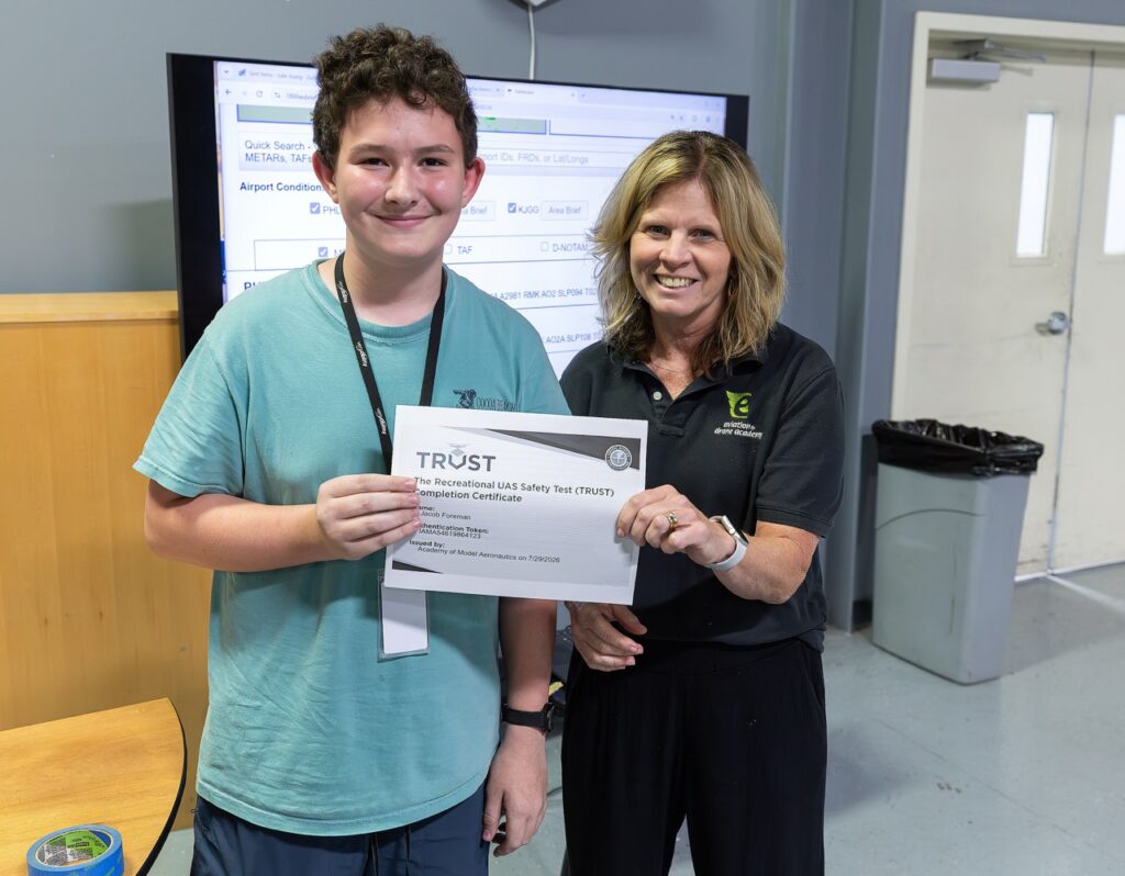 A young person smiles while holding a TRUST certificate next to a woman in a room with a monitor and doors in the background. Both appear happy and are looking at the camera.